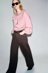 Camisa de manga larga color rosa claro con finas rayas verticales en tono rosado más oscuro. Presenta cuello mao y un gran volante fruncido que recorre el frente, desde los hombros hasta el centro. Los puños son elásticos.