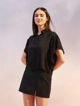 Camisa negra de manga corta con cuello camisero y cierre frontal de botones. Presenta un corte holgado y un dobladillo asimétrico.