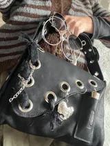 Cartera de cuero negra con correa regulable, estilo bandolera o al hombro. Decorada con charm de corazón metálico, encaje negro, cadenas y aros metálicos.