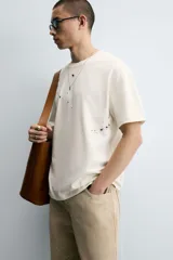 Camiseta color crudo de corte holgado, cuello redondo y manga corta. Tiene estampado de salpicaduras de pintura negra en el frente y texto estampado en la espalda.