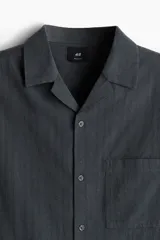 Camisa de manga corta color gris oscuro, confeccionada en mezcla de lino y algodón. Tiene cuello abierto, tapeta lisa, bolsillo de parche en el pecho y bajo recto con aberturas laterales.