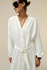Vestido midi blanco de crepe con cuello redondo y escote pico, manga larga con puños y cinturón de la misma tela con hebilla metálica. Cierre frontal con botones metálicos.