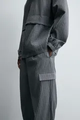 Pantalón cargo gris con textura acanalada, corte relaxed fit, cintura elástica con pliegues frontales, bolsillos laterales tipo cargo con solapa, bolsillos frontales y traseros de vivo, cierre frontal con cremallera y botón.