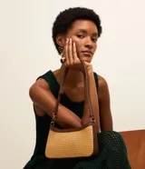 Cartera baguette color beige con efecto paja y detalles en cuero sintético marrón. Tiene correa de hombro ajustable y cierre por cremallera.