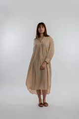 Vestido camisero midi color beige, de corte holgado, con cuello camisero, mangas largas con puños abotonados y aberturas laterales en el ruedo.