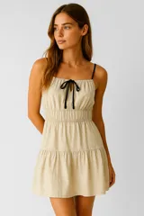 Vestido corto color beige, con breteles finos negros, escote recto y lazo decorativo en el centro del pecho. Tiene corte imperio con falda de dos niveles.
