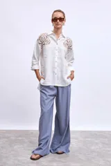 Camisa blanca de lino con cuello clásico, cierre frontal con botones y mangas largas con puños abotonados. Presenta apliques de encaje en hombros y bolsillos delanteros.