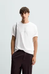 Camiseta blanca de corte holgado, cuello redondo y manga corta. Estampado frontal con texto en color negro y parche en la espalda. Detalle de pliegues en la espalda.
