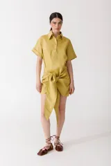 Vestido corto color mostaza, tipo camisa con cuello clásico y mangas cortas. Presenta un nudo en la cintura.