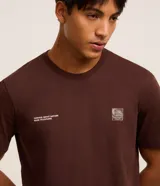 Remera marrón de algodón, corte clásico, con cuello redondo y manga corta. Presenta un pequeño estampado con lettering y un logo en el pecho.