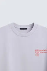 Sudadera lila de corte holgado con cuello redondo y manga corta. Presenta textos estampados en color rojo en el frente y la espalda. Tiene efecto lavado y acabados irregulares.