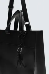 Bolso de hombro negro con compartimento principal con cierre mediante imán y bolsillo interior con cierre. Incluye charm colgante, dos asas de mano y un asa de hombro ajustable y extraíble.