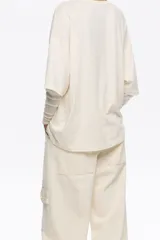 Camiseta oversize de algodón y poliéster color crema con cuello redondo y manga corta ancha. Incluye una manga interior ajustada de tul transparente con estampado sutil. Estampado frontal con diseño de paisaje, árboles, aves y texto.