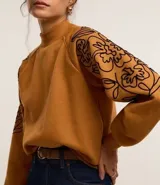 Sweater color ocre con cuello alto y mangas largas con sisas raglán. El sweater tiene bordados de flores en los laterales de las mangas.