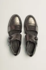 Zapato bajo de cuero negro, estilo monk strap con dos hebillas ajustables sobre el empeine. Presenta detalles de brogue (perforaciones) en la puntera y alrededor del borde superior. Tiene un taco bajo y ancho.