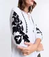 Blusa amplia de viscolino color blanco con bordados florales negros en mangas y escote, lazo en el cuello y mangas 3/4 con puño ajustado.