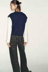 Prenda de manga larga blanca con un chaleco de tejido de punto azul marino superpuesto, creando un efecto de dos piezas. El chaleco es de cuello redondo y corte recto.