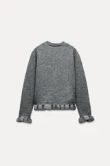 Cárdigan gris jaspeado de punto con cuello redondo y manga larga. Presenta volantes en el borde de los botones, en el bajo y en los puños.