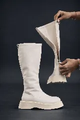 Bota alta tipo bucanera de color blanco hueso, con caña ajustada de material elástico y cierre lateral completo. Posee una plataforma gruesa y dentada en color beige claro.