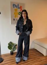 Campera de jean azul oscuro con costuras en contraste color crema, corte cropped, cuello clásico, cierre frontal con botones metálicos plateados, bolsillos de parche en el pecho y mangas largas con puños anchos con dobladillo en color gris. 