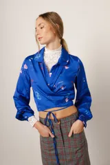Camisa crop azul con estampado de motivos de fiesta, cuello camisero, mangas largas con puños abotonados y lazos para atar en la cintura.