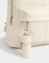 Mochila de cuerina color crudo con bolsillo frontal con cierre y charm decorativo con cierre. Cuenta con correas regulables y manija superior.