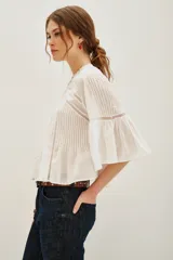 Camisa blanca de algodón con cuello alto y volados, mangas largas abullonadas con puños elásticos. Presenta detalles de encaje y frunces en el frente y la espalda.