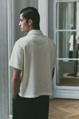 Camisa de manga corta color blanco hueso con textura. Tiene cuello abierto, tapeta lisa y bajo recto.