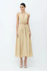 Vestido midi color beige, con cuello halter, falda amplia y corpiño con lazo en el cuello y la espalda. Presenta un detalle de vuelta en la parte delantera, dos botones forrados en el escote y una cremallera invisible en la espalda.