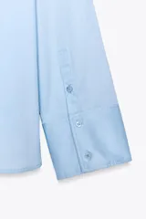 Camisa celeste de popelín con cuello solapa, escote pico y manga larga. Tiene bajo asimétrico con aberturas laterales y cierre frontal con botones.
