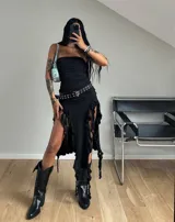 Vestido negro con escote strapless, corte ajustado y falda midi irregular con aberturas laterales y flecos. Se complementa con un cinturón con hebilla metálica y botas texanas negras.
