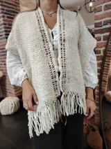 Ruana de lana color crudo tejida a mano, con flecos en el ruedo y detalles de cuero marrón. Diseño abierto y corte holgado.