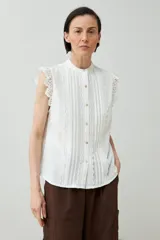 Camisa blanca sin mangas con cuello mao y cierre frontal con botones. Presenta detalles de encaje en los hombros y en el frente.