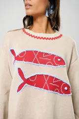 Sweater tejido color crudo con cuello redondo y mangas largas. Presenta dos peces rojos con detalles en azul bordados en el frente. Tiene ribete ondulado rojo en cuello, puños y ruedo.