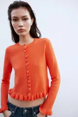 Chaqueta de punto rosa con cuello redondo y manga larga. Detalle de volantes en los bordes y cierre frontal con botones.