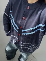 Saco de punto calado color beige claro, con cuello redondo y cierre frontal de botones azules. Presenta detalles decorativos en crochet verde y negro en el frente y puños.