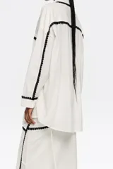 Camisa larga color blanco con borde de encaje negro, de corte recto, con cuello camisero, manga larga con puño y botón, y cierre frontal con botones.