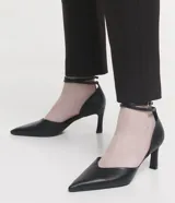 Zapato de tacón tipo scarpin color negro, confeccionado en poliuretano con acabado símil cuero. Presenta puntera fina, talón cerrado con laterales abiertos y una pulsera fina con hebilla dorada que se ajusta al tobillo. El diseño incluye un escote en V en la parte superior y un tacón de 7,5 cm.