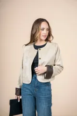 Chaqueta corta color beige, con cuello redondo, cierre frontal con botones dorados, bolsillos de parche y puños en contraste color marrón.