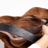 Cartera tipo bucket color marrón, confeccionada en cuero graneado con correa de cuero engrasado y hebilla metálica.
