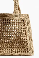 Bolso de mano tipo shopper, confeccionado en paja trenzada color beige, con detalles en cuero marrón. Incluye correa fina extraíble y etiqueta decorativa en un asa.