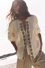 Blusa color crudo de cuello redondo con escote pico y manga larga. Detalle de bordados delanteros y abalorios. Cierre frontal con botones.