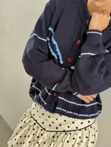 Saco de punto calado color beige claro, con cuello redondo y cierre frontal de botones azules. Presenta detalles decorativos en crochet verde y negro en el frente y puños.