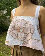 Top de lino blanco con bordado floral de gran tamaño en tonos cobrizos. Presenta un diseño de espalda descubierta con tirantes anchos cruzados y un bajo asimétrico y fluido.