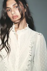 Camisa blanca semitransparente con cuello subido, manga larga con puños, pliegues y volantes combinados. Cierre frontal con botones.