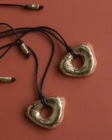 Collar con dije dorado irregular con agujero central, colgando de cordón negro ajustable.