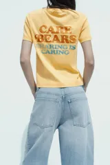Camiseta color amarillo con efecto lavado, cuello redondo y mangas cortas. Estampado frontal con un oso de los Care Bears™ y la frase "free hugs".