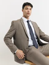 Traje de vestir color marrón topo, compuesto por saco de dos botones con solapa de muesca, dos bolsillos delanteros con solapa y un bolsillo superior. Pantalón de vestir con corte recto y bolsillos laterales. Incluye camisa blanca y corbata azul con lunares blancos.