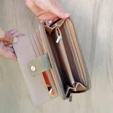 Billetera larga color marrón claro, con cierre de broche a presión en la solapa frontal y cierre de cremallera en el borde lateral. Presenta múltiples compartimentos internos y un bolsillo exterior con cierre.