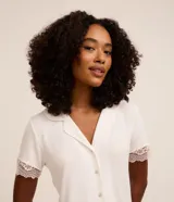 Conjunto de pijama femenino confeccionado en malla de viscosa y elastano, modelo short doll americano, compuesto por una camisa abotonada con solapa y un short con cintura elástica. Presenta un estampado de lunares y detalles de encaje en las mangas y el ruedo del short.